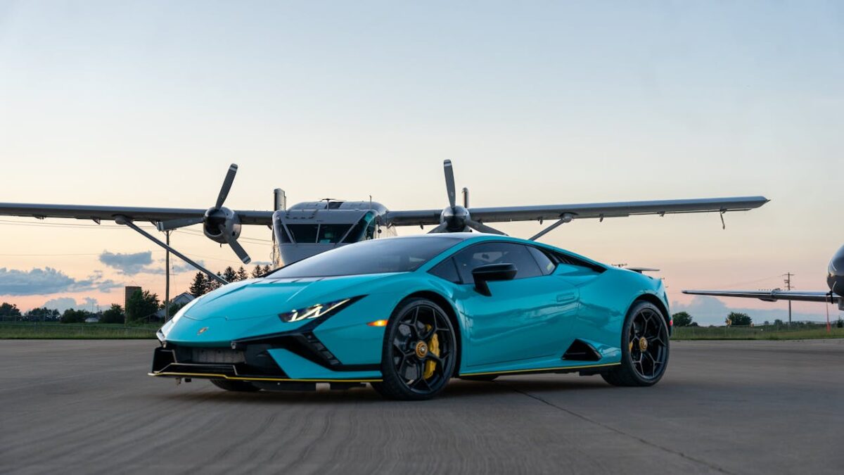 A stylish turquoise sports car parked in front of an airplane at sunset.